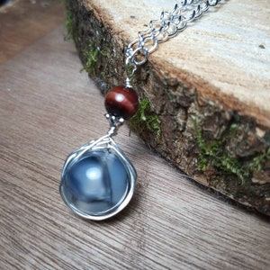 May include: A silver chain necklace with a grey agate stone pendant wrapped in silver wire. The pendant is accented with a small red tiger's eye bead.
