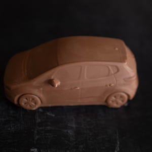 May include: A brown miniature car model, featuring a hatchback design with visible wheels, side mirrors, and door handles. The car has a rounded roof and a smooth finish. The model is set against a dark background.