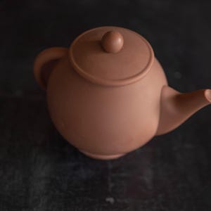 May include: A terracotta teapot with a rounded body and a lid with a small knob on top. The teapot has a spout on one side and a handle on the other.