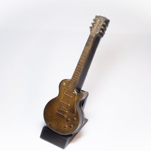 May include: A gold and black electric guitar candle on a black stand. The guitar has a detailed design and is a realistic representation of a real guitar.