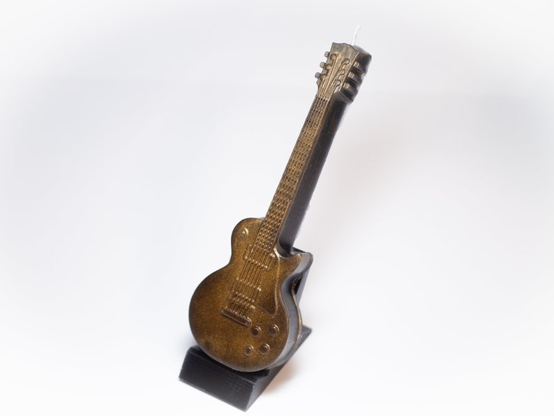 May include: A gold and black electric guitar candle on a black stand. The guitar has a detailed design and is a realistic representation of a real guitar.