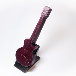 May include: A dark red electric guitar shaped candle with a black stand. The candle is detailed with a textured body and a white wick.