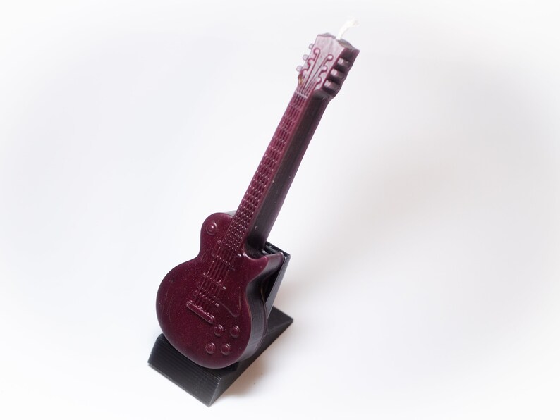 May include: A dark red electric guitar shaped candle with a black stand. The candle is detailed with a textured body and a white wick.