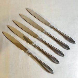 May include: Five stainless steel dinner knives arranged diagonally. Each knife has a long, slender blade and a handle with a slight curve. The knives are set against a plain, off-white background.