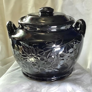 May include: A glossy black ceramic bean pot with a matching lid and two side handles. The exterior surface features a raised, embossed pattern of leaves and berries that wraps around the body of the round, wide-mouthed vessel.