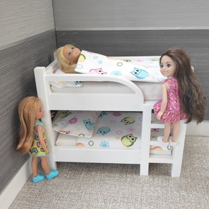May include: White wooden bunk bed with a white, pink, and blue owl patterned bedding. Two dolls are on the bed, one is sleeping and the other is standing on the ladder.