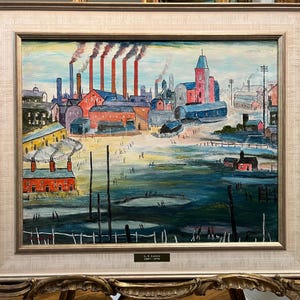 May include: An oil painting in a gold frame depicts an industrial town with red brick buildings, smokestacks, and figures. The sky is blue with wisps of smoke. The painting is signed "L.S. Lowry 1887-1976".