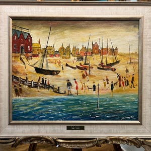 May include: An oil painting depicting a seaside scene with a beach, boats, and figures. The artwork features a red building, boats with masts, and people strolling along the shore. The painting is framed in a silver and gold ornate frame.