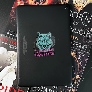 May include: Black tablet with a sticker featuring a teal wolf design and the text "I LIKE MY MEN FERAL & FATED." The tablet is surrounded by books with titles like "BORN BY NLIGHT" and "ALI HA."
