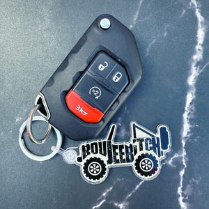 May include: Black car key fob with red accents and a silver keyring. A clear acrylic keychain in the shape of a Jeep with the text "BOUJEEBITCH" in black and white is attached to the keyring.