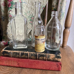 Op de afbeelding: Vier vintage glazen flessen, twee helder en twee met etiketten. De flessen zijn gerangschikt op twee boeken, een zwart en een rood. Het etiket op een flesje luidt "60 Drac. Chicine - Aspirin Houd. DOS: 1 a 3 tps. FORMULE:  Laboratoires Houd. 57 Rue D'Anvers, Paris"