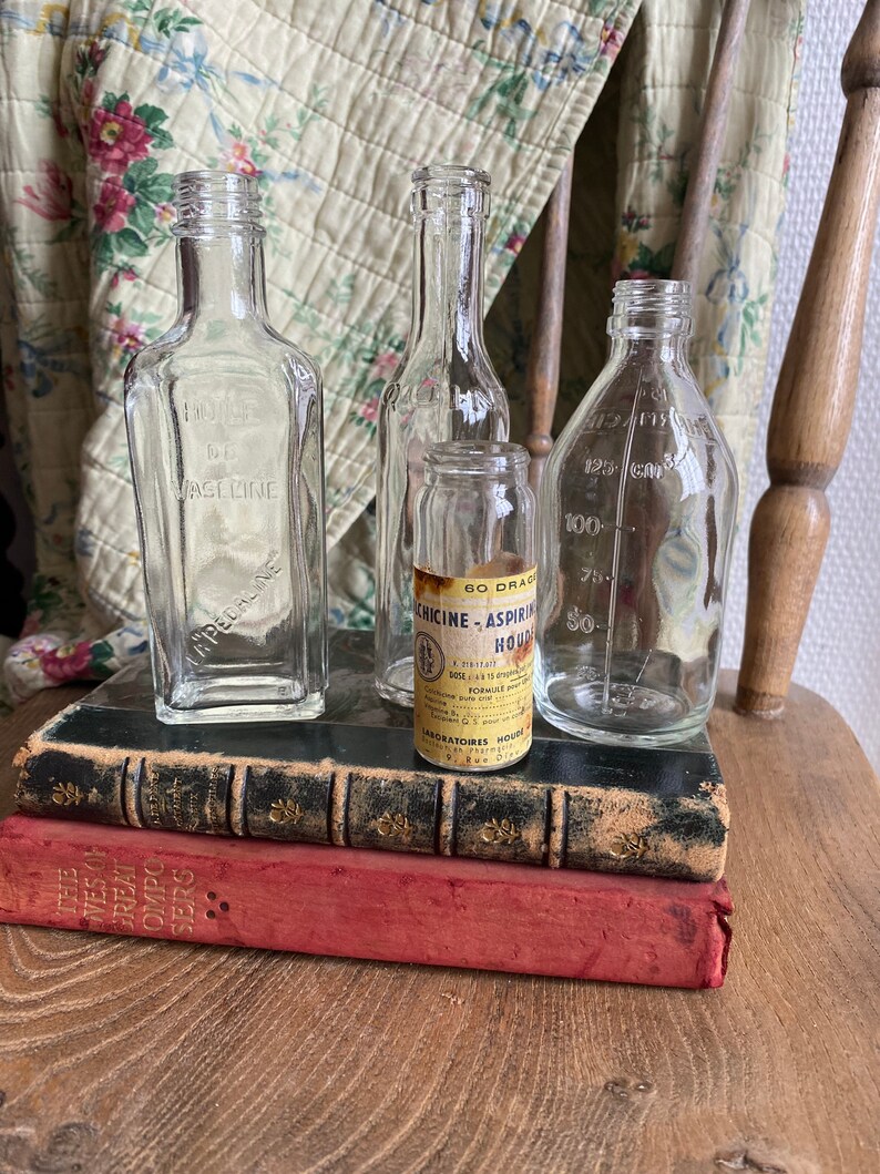 Op de afbeelding: Vier vintage glazen flessen, twee helder en twee met etiketten. De flessen zijn gerangschikt op twee boeken, een zwart en een rood. Het etiket op een flesje luidt "60 Drac. Chicine - Aspirin Houd. DOS: 1 a 3 tps. FORMULE:  Laboratoires Houd. 57 Rue D'Anvers, Paris"