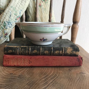 May include: A white ceramic gravy boat with a green and gold trim. The boat is sitting on top of two antique books. The books are on a wooden surface.