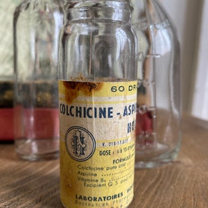 Op de afbeelding: Een vintage glazen fles met een geel etiket waarop "Colchicine-Aspirine HO" staat. Het etiket bevat ook de tekst "V. 218-17.077", "DOSE : 4 a 15 drops", "FORMULE", "Colchicine pure crist", "Aspirine", "Vitamine B1", "Excipient Q. S. pour", "LABORATOIRES HO", "Docteur en Pharmacie", "9, Rue Di".