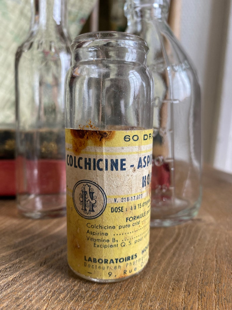 Op de afbeelding: Een vintage glazen fles met een geel etiket waarop "Colchicine-Aspirine HO" staat. Het etiket bevat ook de tekst "V. 218-17.077", "DOSE : 4 a 15 drops", "FORMULE", "Colchicine pure crist", "Aspirine", "Vitamine B1", "Excipient Q. S. pour", "LABORATOIRES HO", "Docteur en Pharmacie", "9, Rue Di".