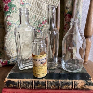Op de afbeelding: Vier vintage glazen flessen met etiketten, die op een stapel oude boeken staan. De flessen zijn van helder glas en hebben verschillende vormen en maten. De etiketten zijn in het Frans en bevatten de tekst "Huile de Vaseline" en "M&eacute;dicine-Aspirine Houde".