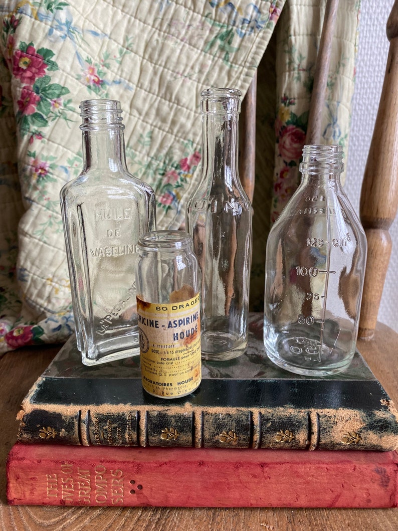 Op de afbeelding: Vier vintage glazen flessen met etiketten, die op een stapel oude boeken staan. De flessen zijn van helder glas en hebben verschillende vormen en maten. De etiketten zijn in het Frans en bevatten de tekst "Huile de Vaseline" en "M&eacute;dicine-Aspirine Houde".