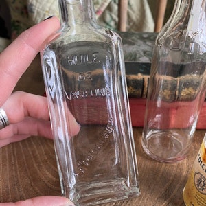 Op de afbeelding: Twee doorzichtige glazen flessen met een vierkante vorm. De fles aan de linkerkant heeft de tekst "HUILE DE VASELINE" op de voorkant gedrukt. De fles aan de rechterkant heeft een geel etiket met tekst in een vreemde taal.