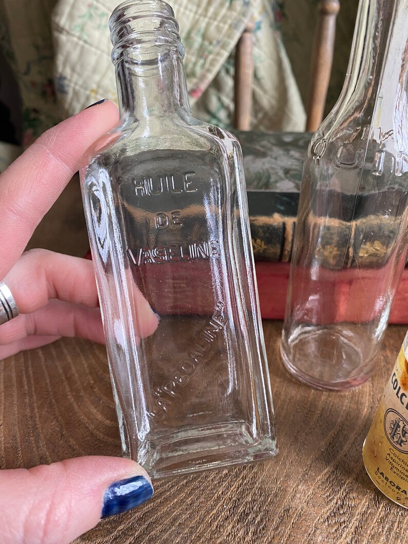 Op de afbeelding: Twee doorzichtige glazen flessen met een vierkante vorm. De fles aan de linkerkant heeft de tekst "HUILE DE VASELINE" op de voorkant gedrukt. De fles aan de rechterkant heeft een geel etiket met tekst in een vreemde taal.