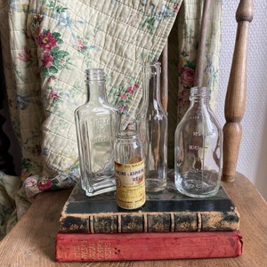 Op de afbeelding: Vier vintage glazen flessen met etiketten, die op een stapel van drie boeken staan. De flessen zijn helder en hebben verschillende vormen en maten. De boeken zijn gebonden in zwart leer en rode stof. De flessen staan op een houten oppervlak.
