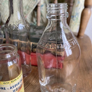 Op de afbeelding: Drie heldere glazen flessen met een vintage uitstraling. De fles in het midden heeft het woord "PHARMACIE" gegraveerd op de voorkant. De fles links heeft een etiket met de tekst "60 DRAGEES NE-ASPIRINE HOUDE" en andere tekst in het Frans.
