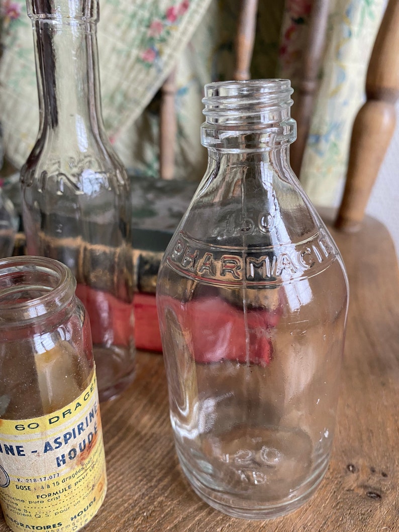 Op de afbeelding: Drie heldere glazen flessen met een vintage uitstraling. De fles in het midden heeft het woord "PHARMACIE" gegraveerd op de voorkant. De fles links heeft een etiket met de tekst "60 DRAGEES NE-ASPIRINE HOUDE" en andere tekst in het Frans.