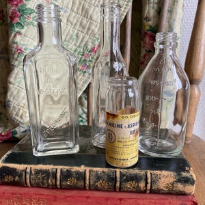 Op de afbeelding: Vier vintage glazen flessen met etiketten, die op twee antieke boeken staan. De flessen zijn van helder glas en hebben verschillende vormen en maten. De etiketten zijn in het Frans en Engels. De boeken zijn gebonden in leer en hebben gouden letters.