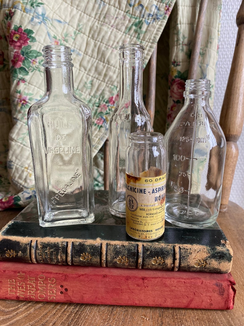 Op de afbeelding: Vier vintage glazen flessen met etiketten, die op twee antieke boeken staan. De flessen zijn van helder glas en hebben verschillende vormen en maten. De etiketten zijn in het Frans en Engels. De boeken zijn gebonden in leer en hebben gouden letters.