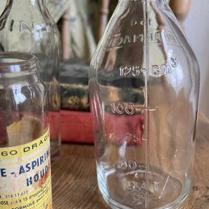 Op de afbeelding: Drie glazen flessen, twee met heldere etiketten en een met een geel etiket waarop "60 DRAGEE - ASPIRIN HOUDE" staat. De fles rechts heeft markeringen voor 125, 100, 75, 50 en 25 kubieke centimeter.