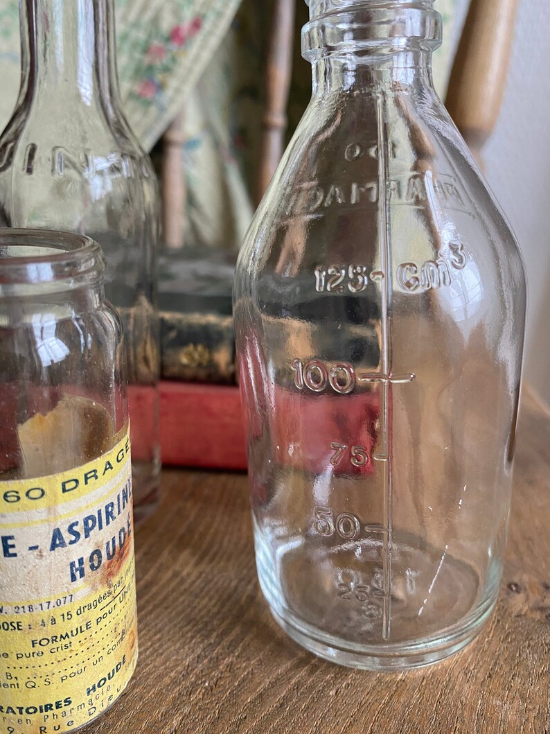 Op de afbeelding: Drie glazen flessen, twee met heldere etiketten en een met een geel etiket waarop "60 DRAGEE - ASPIRIN HOUDE" staat. De fles rechts heeft markeringen voor 125, 100, 75, 50 en 25 kubieke centimeter.