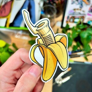 Op de afbeelding: Een gestanste sticker met een gele banaan met een goudkleurig saxofoonmondstuk dat uit de geschilde vrucht komt. De sticker heeft een witte rand en wordt in een hand gehouden.