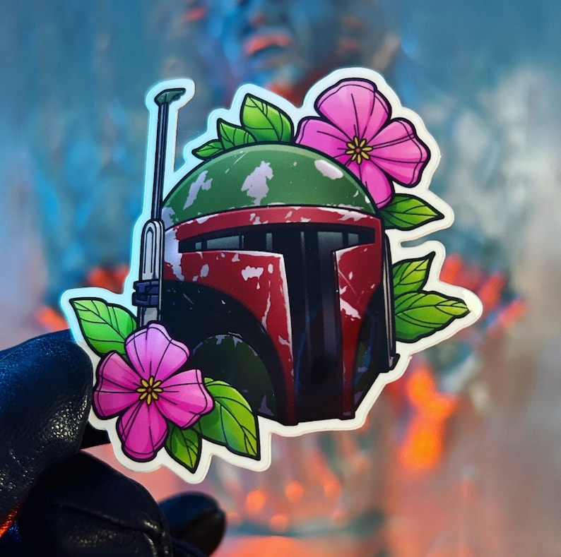May include: A sticker featuring a green and red Star Wars Boba Fett helmet with pink flowers. The helmet has a weathered look and is surrounded by green leaves.
