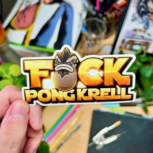 以下が含まれることがあります： 金と白で「F*CK PONGKRELL」の文字が入ったステッカー。漫画のキャラクターが描かれています。ステッカーは手で持たれており、背景には他の画材や絵が見えます。