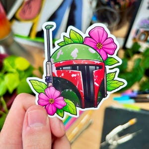 May include: A sticker featuring a green, red, and black Boba Fett helmet with pink flowers and green leaves.