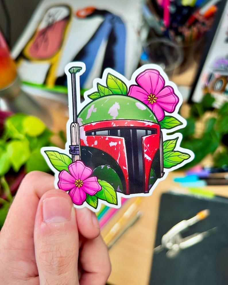 May include: A sticker featuring a green, red, and black Boba Fett helmet with pink flowers and green leaves.