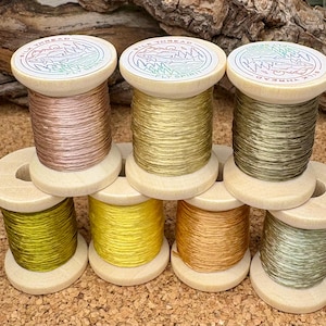 Puede incluir: Una colección de siete carretes de hilo de seda en varios colores, incluyendo rosa, dorado, verde oliva, amarillo y naranja. Cada carrete tiene una base de madera y una parte superior con un diseño circular y el texto "SILK THREAD FLY SPIRIT".