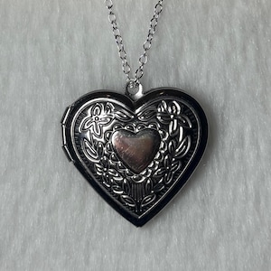 May include: Silver heart-shaped locket necklace. The locket has an ornate floral design around the edges and a smaller heart in the center. The necklace chain is also silver.