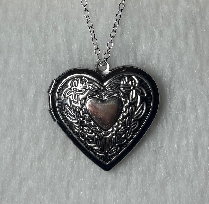 May include: Silver heart-shaped locket necklace. The locket has an ornate floral design around the edges and a smaller heart in the center. The necklace chain is also silver.