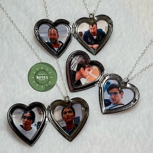 May include: Heart-shaped locket necklaces with silver chains. Each locket is open, revealing a photograph. The lockets are arranged on a white surface. The image also includes a green circular logo with the text "KOTA'S SHOPPE".