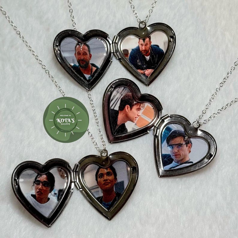 May include: Heart-shaped locket necklaces with silver chains. Each locket is open, revealing a photograph. The lockets are arranged on a white surface. The image also includes a green circular logo with the text "KOTA'S SHOPPE".