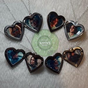 May include: Heart-shaped lockets with silver chains. Each locket contains a small image of a person. The lockets are open and closed, revealing the images inside. The background includes a green circle with the text "WELCOME TO KOTA'S SHOPPE."