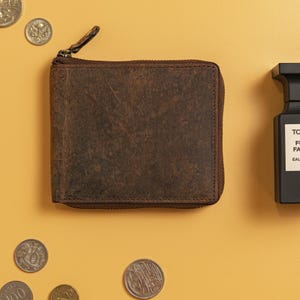 May include: A square brown leather wallet with a dark brown zip closure along the top and side. The leather has a distressed, textured finish with visible grain and natural markings. A small metal zip pull is attached to the top corner.