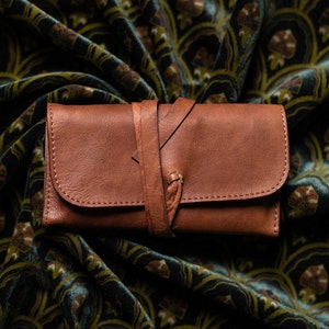 May include: A brown leather wallet with a strap closure, lying on a patterned fabric background.