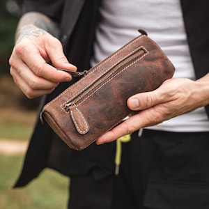 May include: A brown leather pencil case with a zipper closure. The case has a distressed look and is made of high-quality leather.