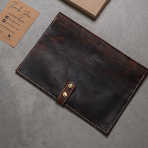 May include: A dark brown leather laptop sleeve with a flap closure and two brass button accents.