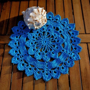 Pode incluir: Uma toalha de mesa de crochê azul brilhante, feita à mão, com uma borda ondulada e um intrincado design floral. Uma grande concha marinha creme repousa sobre a toalha de mesa, criando um tema costeiro. A toalha de mesa é exibida em uma superfície de madeira.