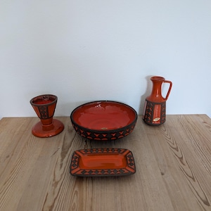 Op de afbeelding: Een verzameling oranje keramiek serviesgoed met zwarte geometrische ontwerpen. De set bevat een kom, een kan, een rechthoekige schaal en een kleine voetbeker. De items zijn geplaatst op een houten oppervlak.