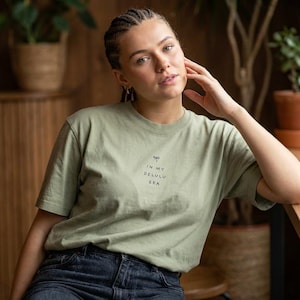 以下が含まれることがあります： 黒色の「IN MY DELULU ERA」という文字が入ったセージグリーンのTシャツ。Tシャツはクルーネックで半袖です。モデルは濃い青色のジーンズを着用しています。