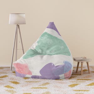 May include: A large, triangular beanbag chair with a white background and abstract shapes in pastel colors. The shapes include pink, purple, blue, and green. The beanbag is on a patterned rug.