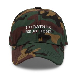 May include: Camouflage baseball cap with the embroidered text "I'D RATHER BE AT HOME" in white. The cap features a curved brim and a black button on top. The fabric has a pattern of green, brown, and tan.